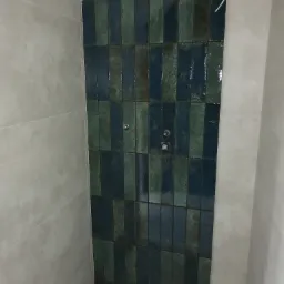 Fragment ściany łazienki wyłożonej pionowymi, błyszczącymi płytkami w odcieniach zieleni i granatu, z widoczną instalacją wodną i sąsiednią ścianą wykończoną szarymi płytami.