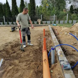 Pracownik z łopatą obok wykopu z ułożonymi rurami kanalizacyjnymi i fundamentami, w tle zagęszczarka spalinowa.