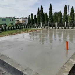 Wylana, mokra płyta fundamentowa z wystającymi rurami kanalizacyjnymi na działce z widokiem na dom i tuje.