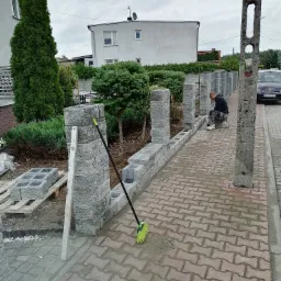 Budowa ogrodzenia z szarych bloczków betonowych, widoczny pracownik układający bloczki, poziomica oparta o słupek, kostka brukowa na chodniku, dom w tle.