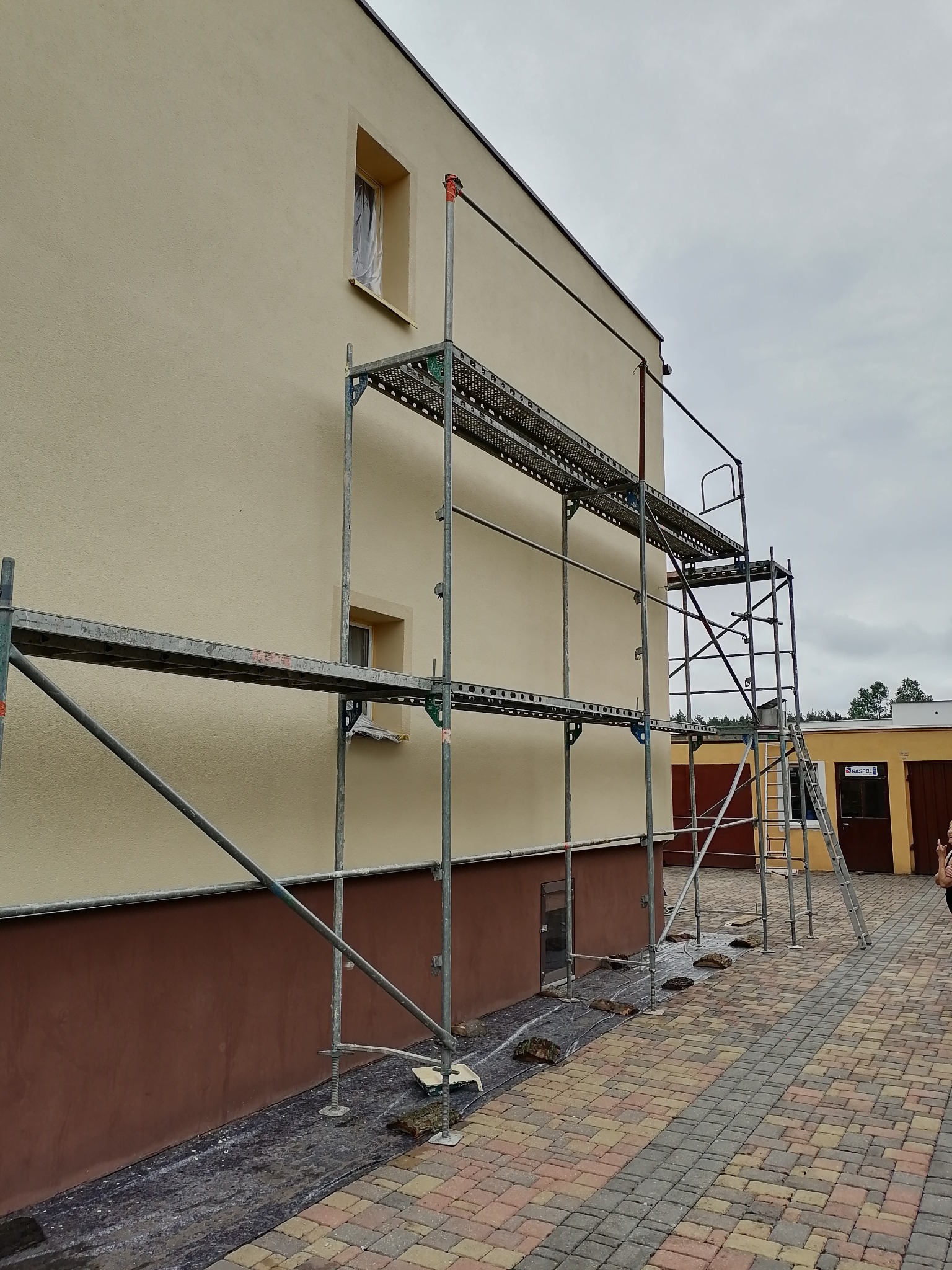 Budynek z beżową elewacją w trakcie prac, widoczne rusztowanie, okna zabezpieczone folią, dolna część elewacji w brązowym kolorze, kostka brukowa na podwórzu.