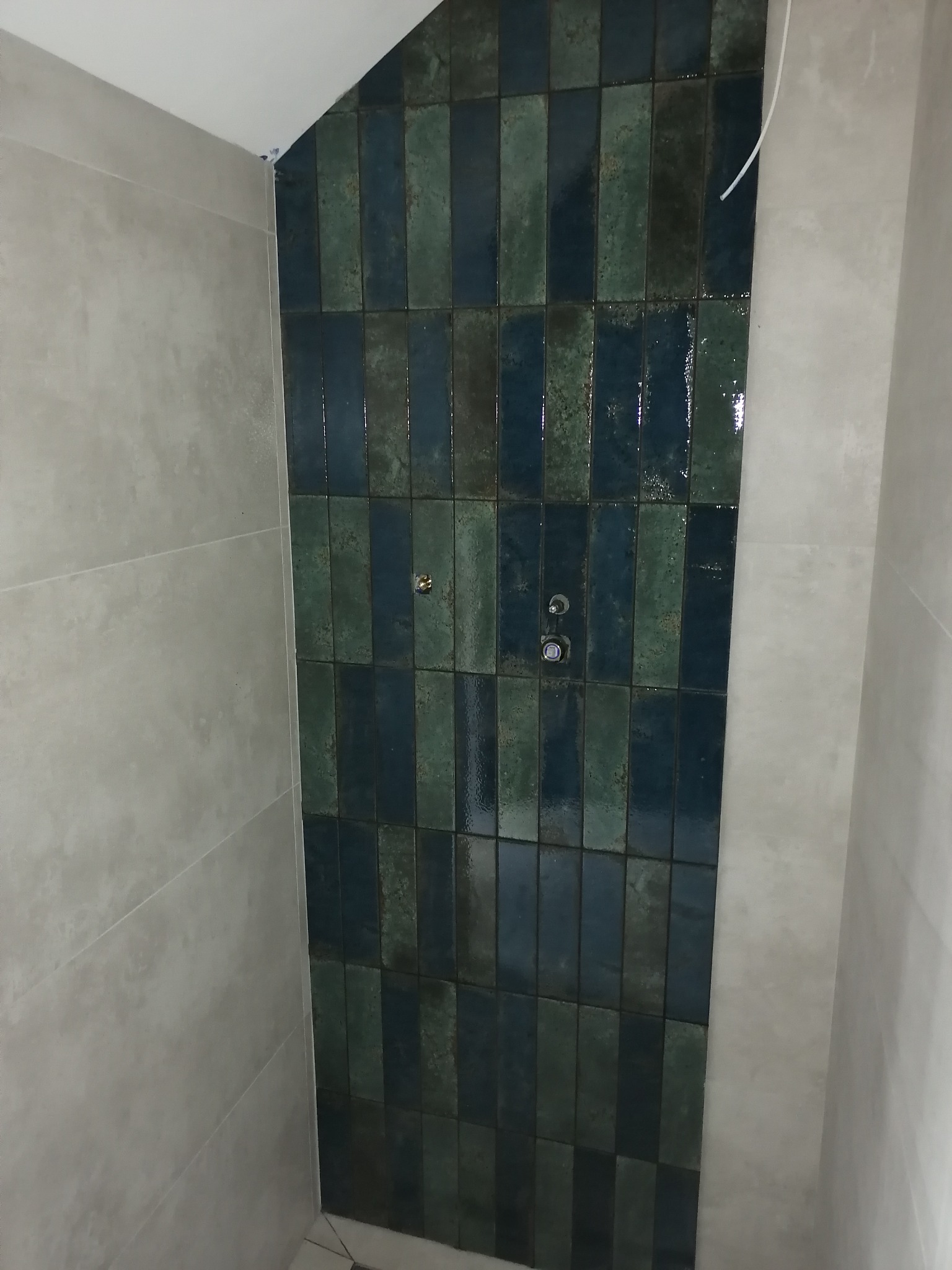 Fragment ściany łazienki wyłożonej pionowymi, błyszczącymi płytkami w odcieniach zieleni i granatu, z widoczną instalacją wodną i sąsiednią ścianą wykończoną szarymi płytami.