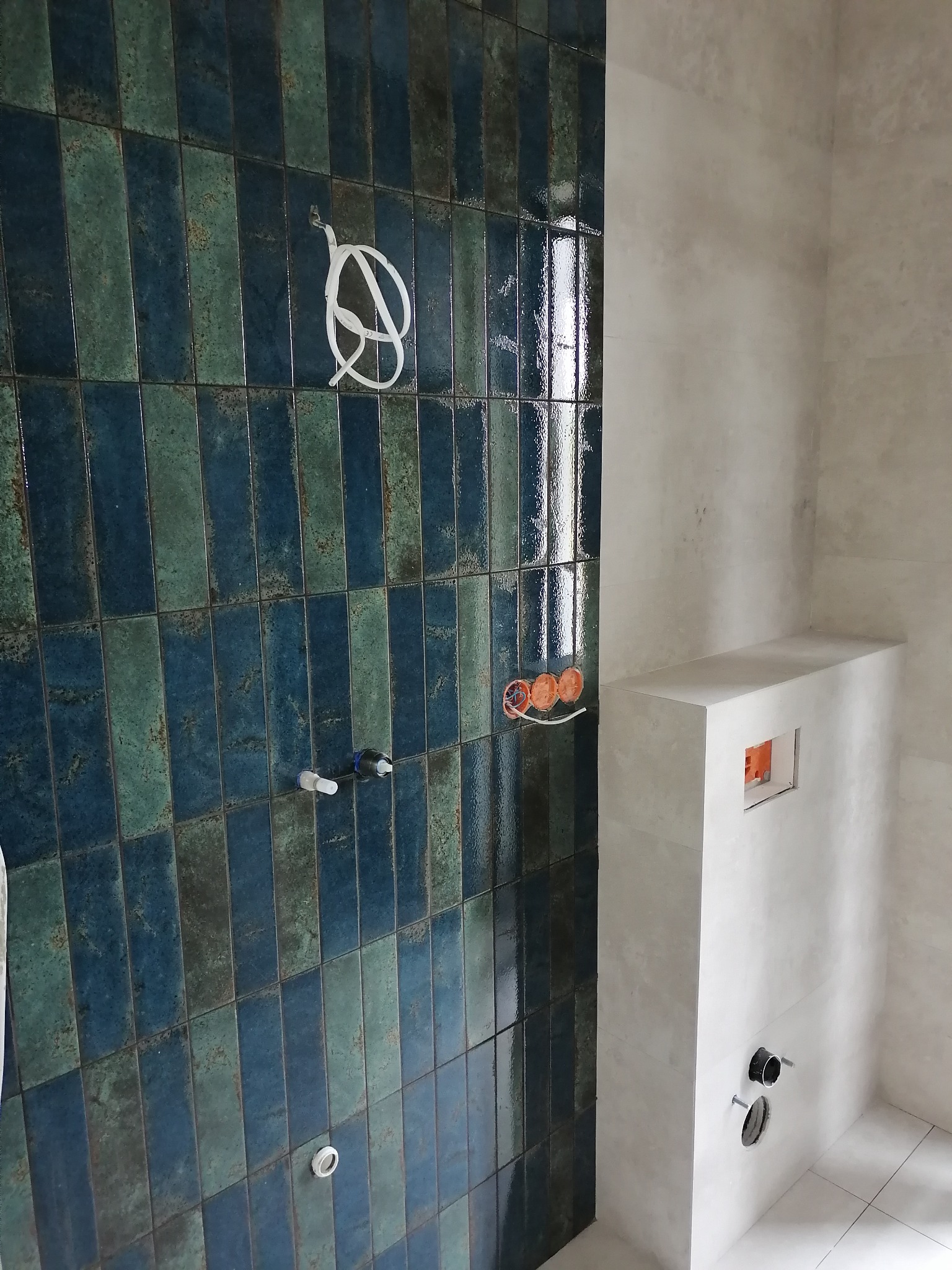 Ściana łazienki z pionowo ułożonymi, błyszczącymi, niebiesko-zielonymi płytkami, widoczne przygotowane instalacje wodno-kanalizacyjne i elektryczne, surowy stan wykończenia.