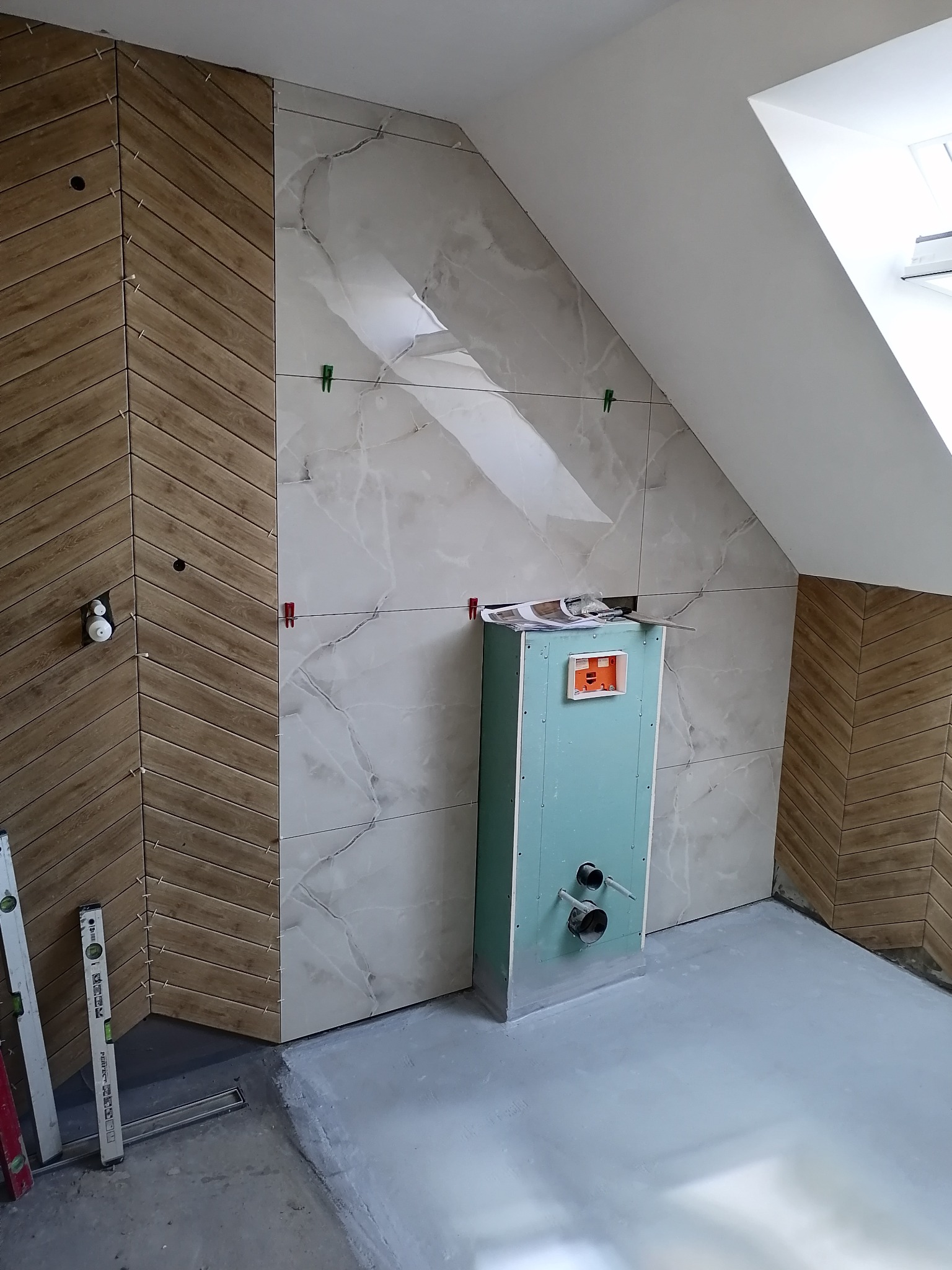 Montaż płytek w łazience na poddaszu: połączenie płytek imitujących drewno ułożonych w jodełkę z marmurowymi płytkami, widoczna obudowa instalacji wodnej i poziomice.