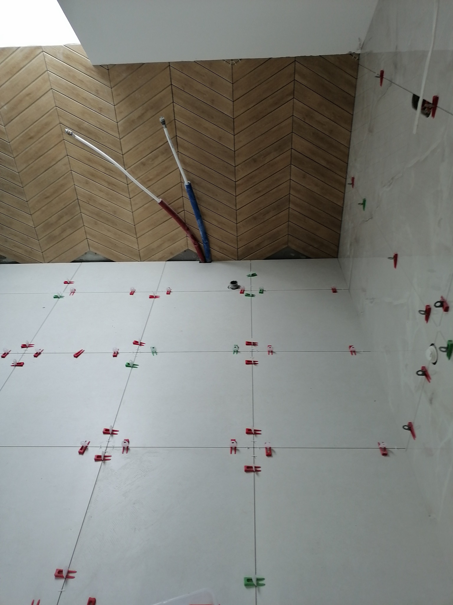 Układanie białych płytek podłogowych z użyciem klipsów poziomujących w łazience w Trzciance, widoczna ściana z brązową okładziną ułożoną we wzór jodełki oraz wystające rurki instalacji.