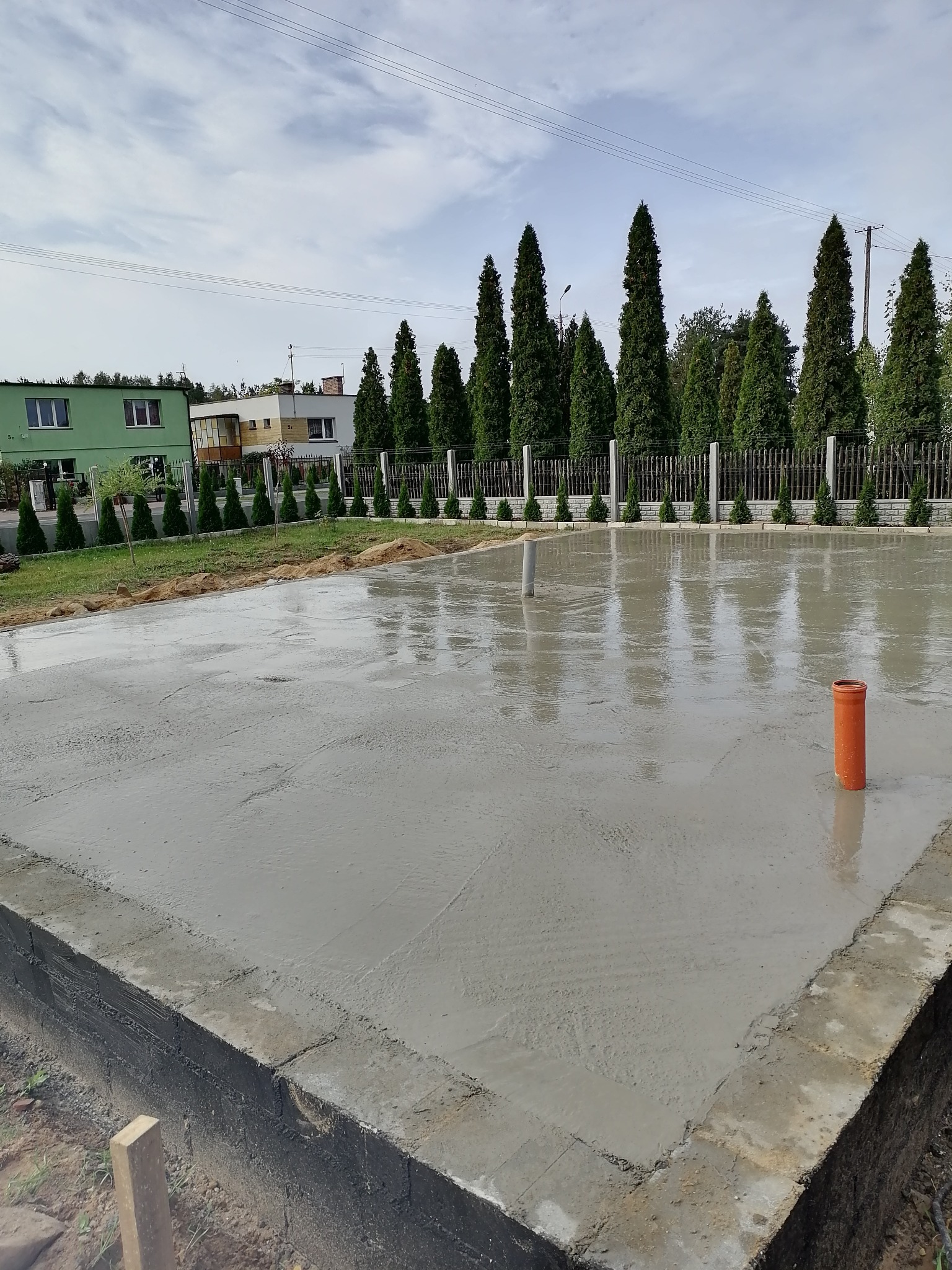 Wylana, mokra płyta fundamentowa z wystającymi rurami kanalizacyjnymi na działce z widokiem na dom i tuje.