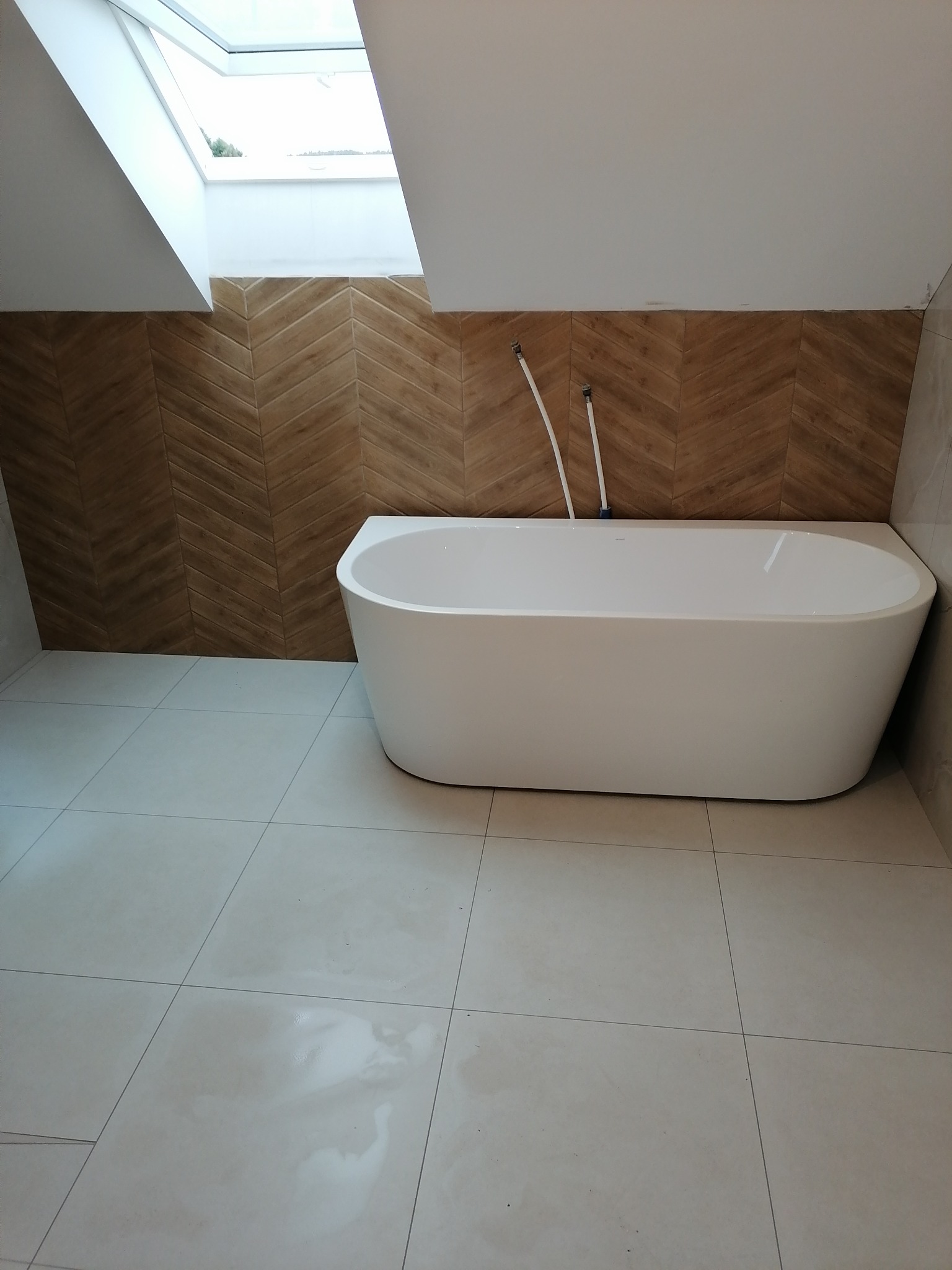 Nowoczesna biała wanna wolnostojąca w łazience na poddaszu z drewnopodobną ścianą ułożoną w jodełkę i oknem dachowym.