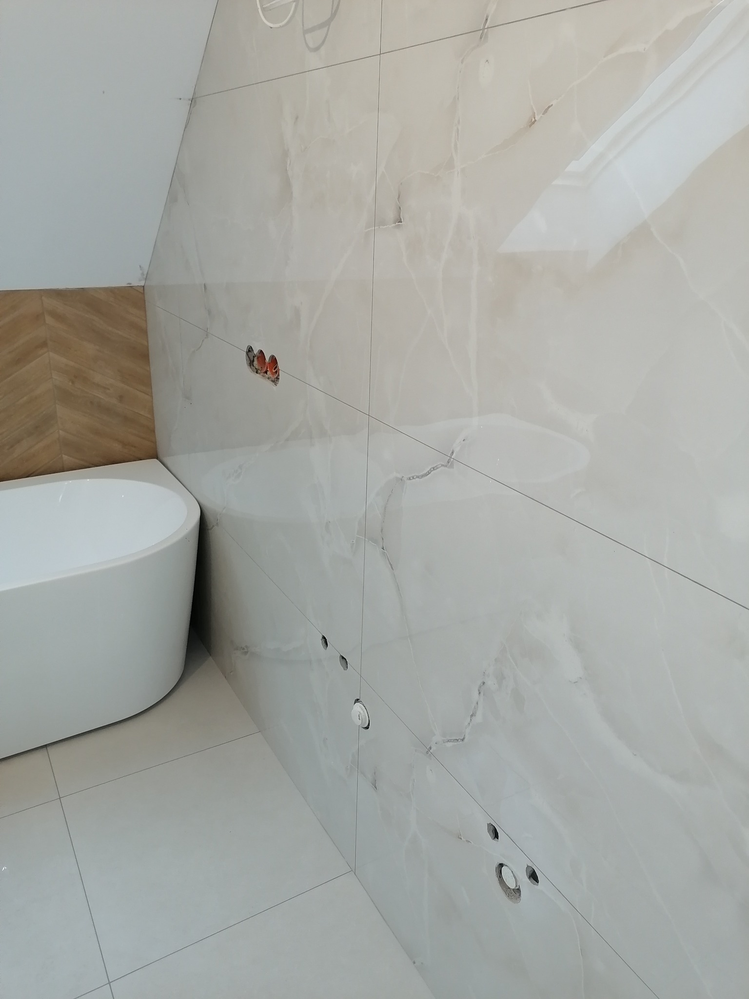 Biała wolnostojąca wanna przy ścianie z płytek imitujących marmur w łazience na poddaszu, widoczne przygotowane punkty instalacji.
