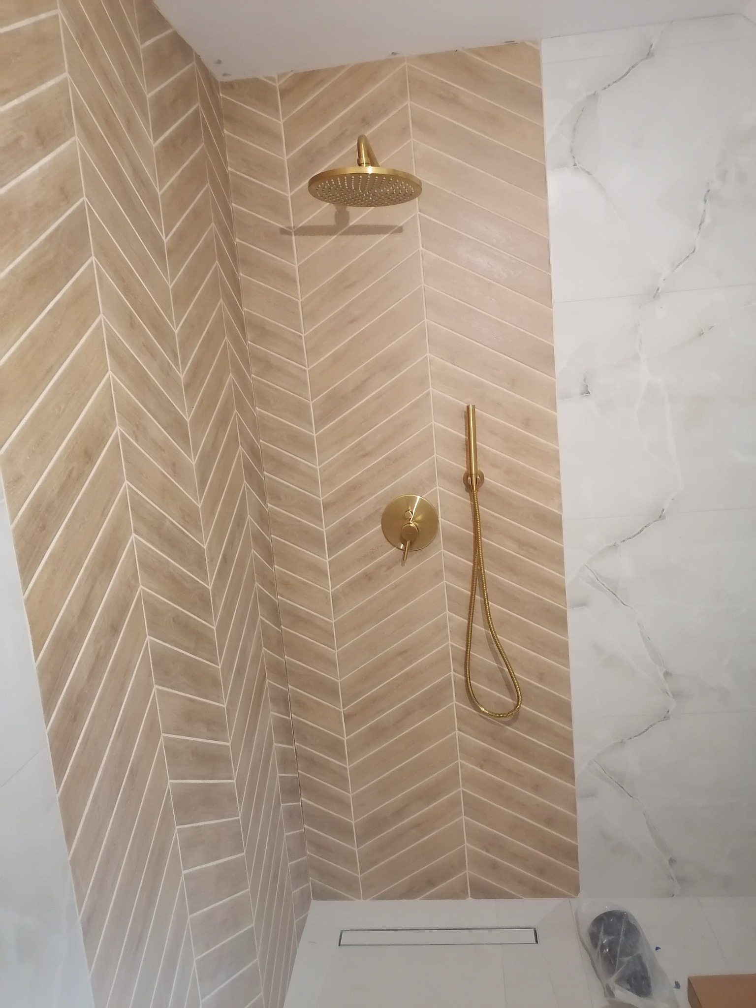 Nowoczesna kabina prysznicowa z beżowymi płytkami ułożonymi w jodełkę i złotą armaturą, kontrastująca z białymi płytami imitującymi marmur.