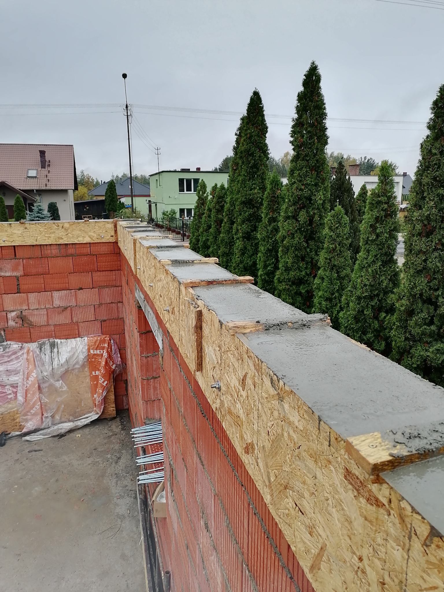 Czerwona ściana z pustaków ceramicznych z szalunkiem i wylewką betonową na górze, widok z góry na plac budowy z tujami w tle.