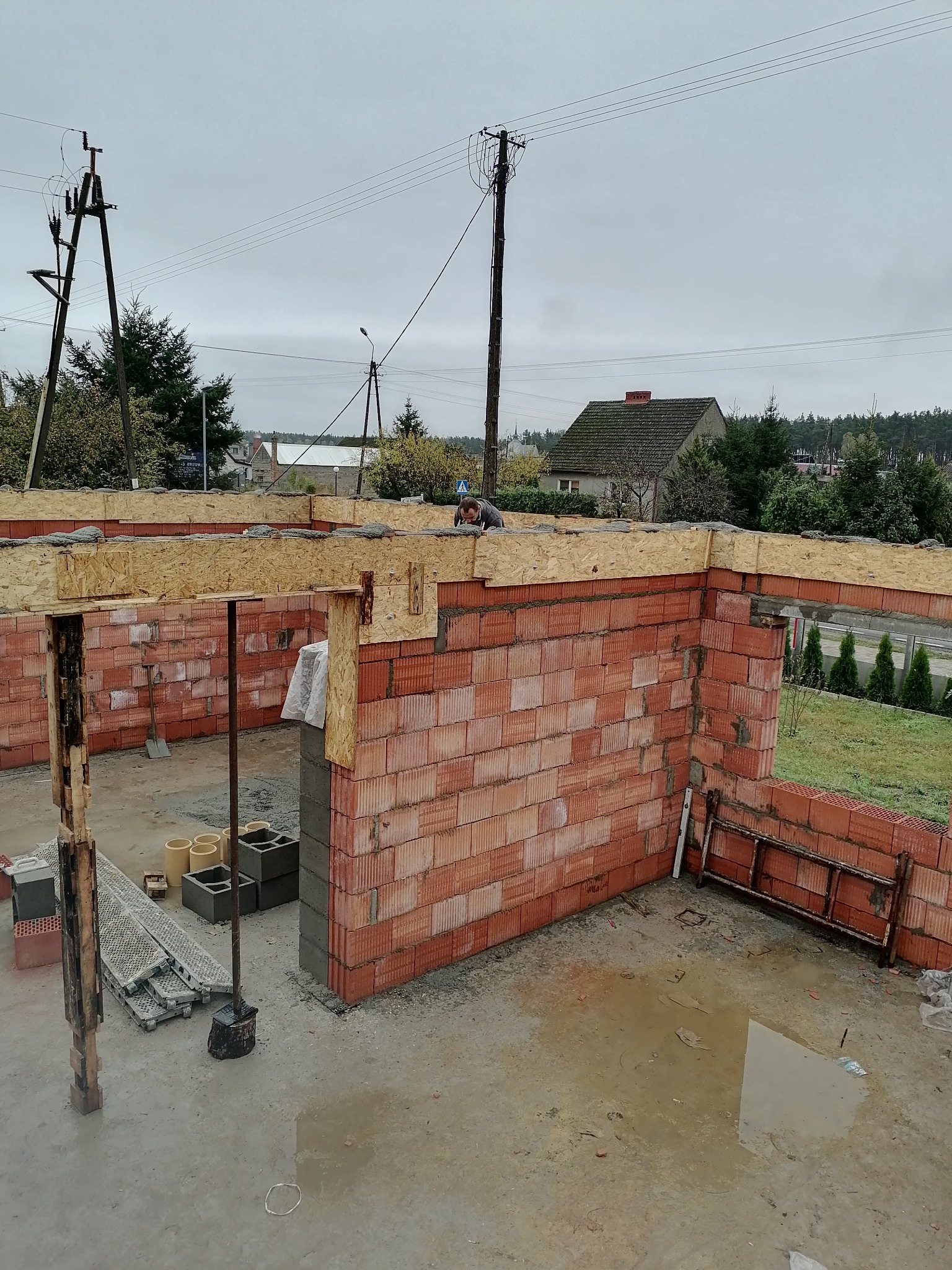 Budowa domu: ściany z czerwonej cegły silikatowej z nadprożami z drewna OSB, widoczny murarz na górze ściany, teren budowy z kałużami i materiałami budowlanymi.