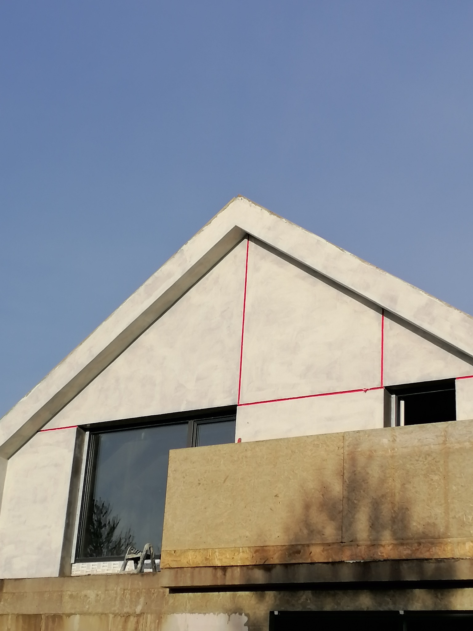 Fragment elewacji domu w trakcie prac wykończeniowych, widoczne tynkowanie, okno z odbiciem drzewa oraz czerwone linie wyznaczające podział elewacji.