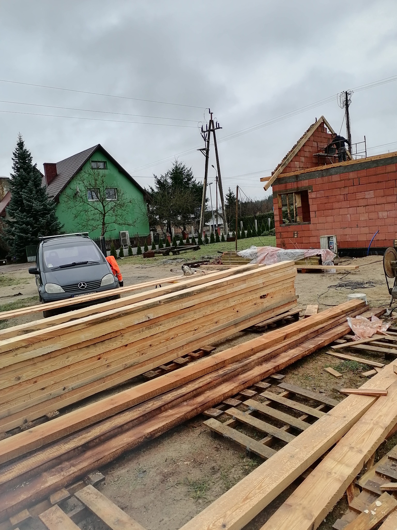 Wznoszenie murów domu z czerwonej cegły, widoczne drewniane elementy konstrukcyjne dachu i pracownik na rusztowaniu, na pierwszym planie stos drewna budowlanego, w tle zielony dom i zaparkowany...