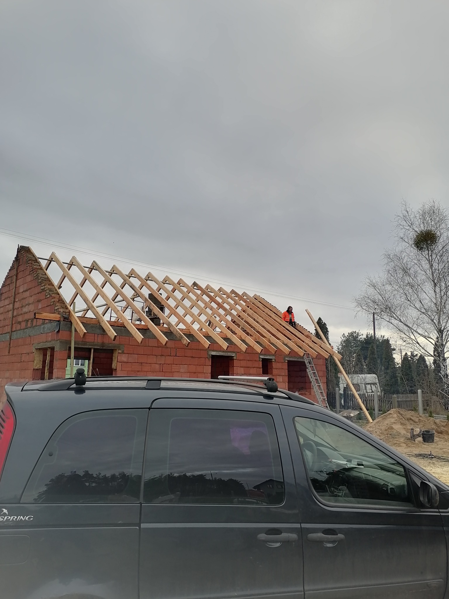 Budowa domu murowanego z widoczną konstrukcją dachu i pracownikiem w pomarańczowej kamizelce, częściowo zasłonięta przez zaparkowany samochód.
