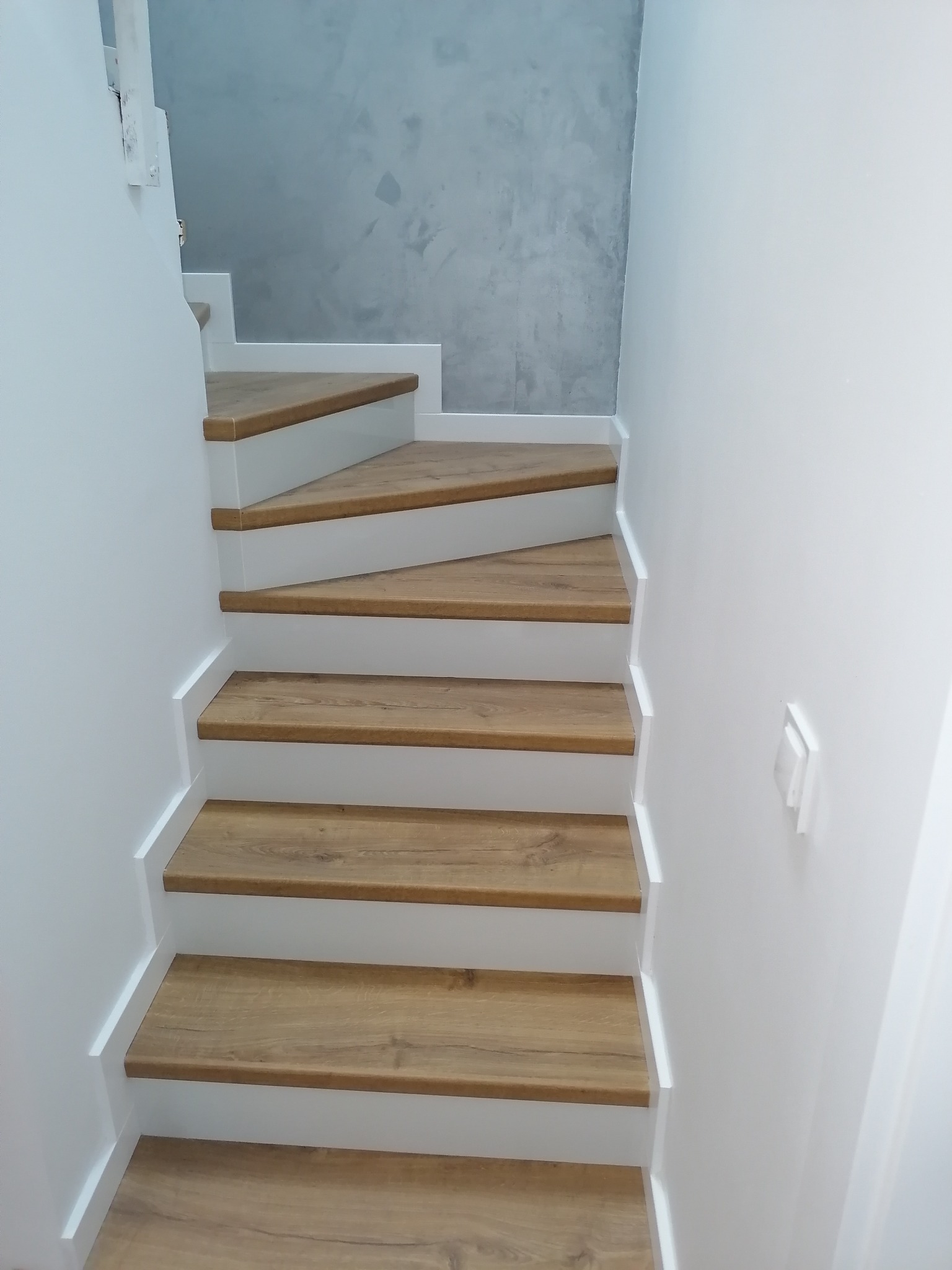 Nowoczesne schody z drewnianymi stopniami i białymi podstopnicami, ściana pomalowana na biało z elementami szarości w tle, minimalistyczny wyłącznik światła po prawej stronie.