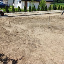 Firma Agpol - Przygotowanie terenu pod budowę drogi: wyrównana ziemia, wbite kołki wyznaczające granice, taczka w tle, widoczny fragment chodnika z kostki i pracownik w oddali.
