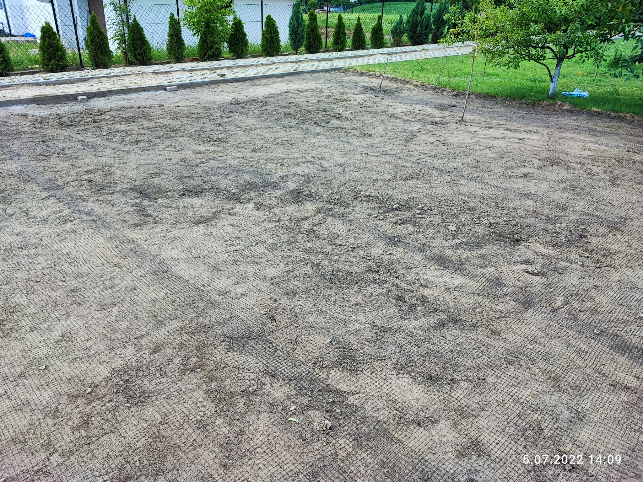 Przygotowany teren pod budowę, pokryty siatką stabilizującą grunt, z widoczną kostką obrzeżową i młodymi drzewami w tle.