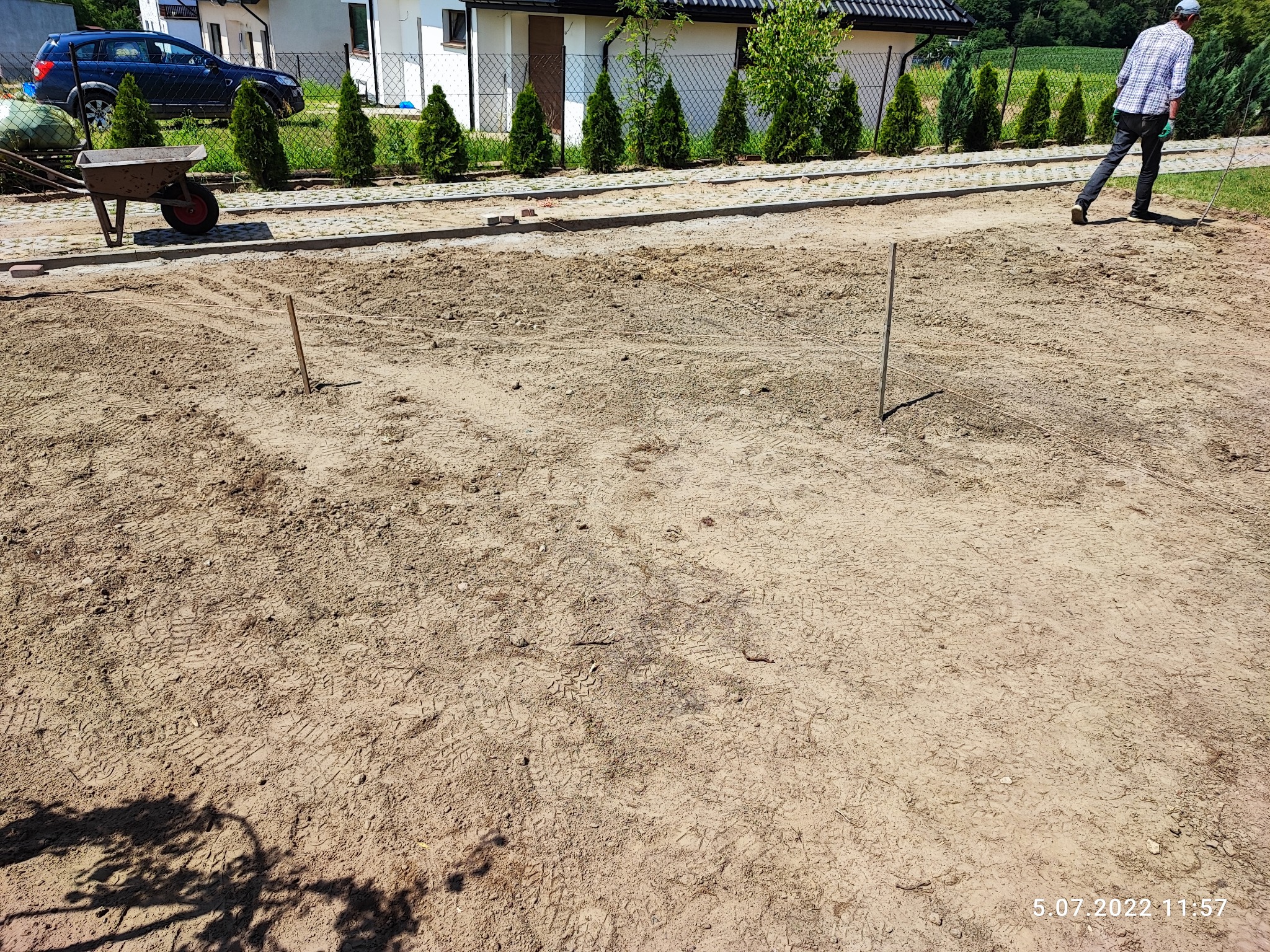 Przygotowanie terenu pod budowę drogi: wyrównana ziemia, wbite kołki wyznaczające granice, taczka w tle, widoczny fragment chodnika z kostki i pracownik w oddali.