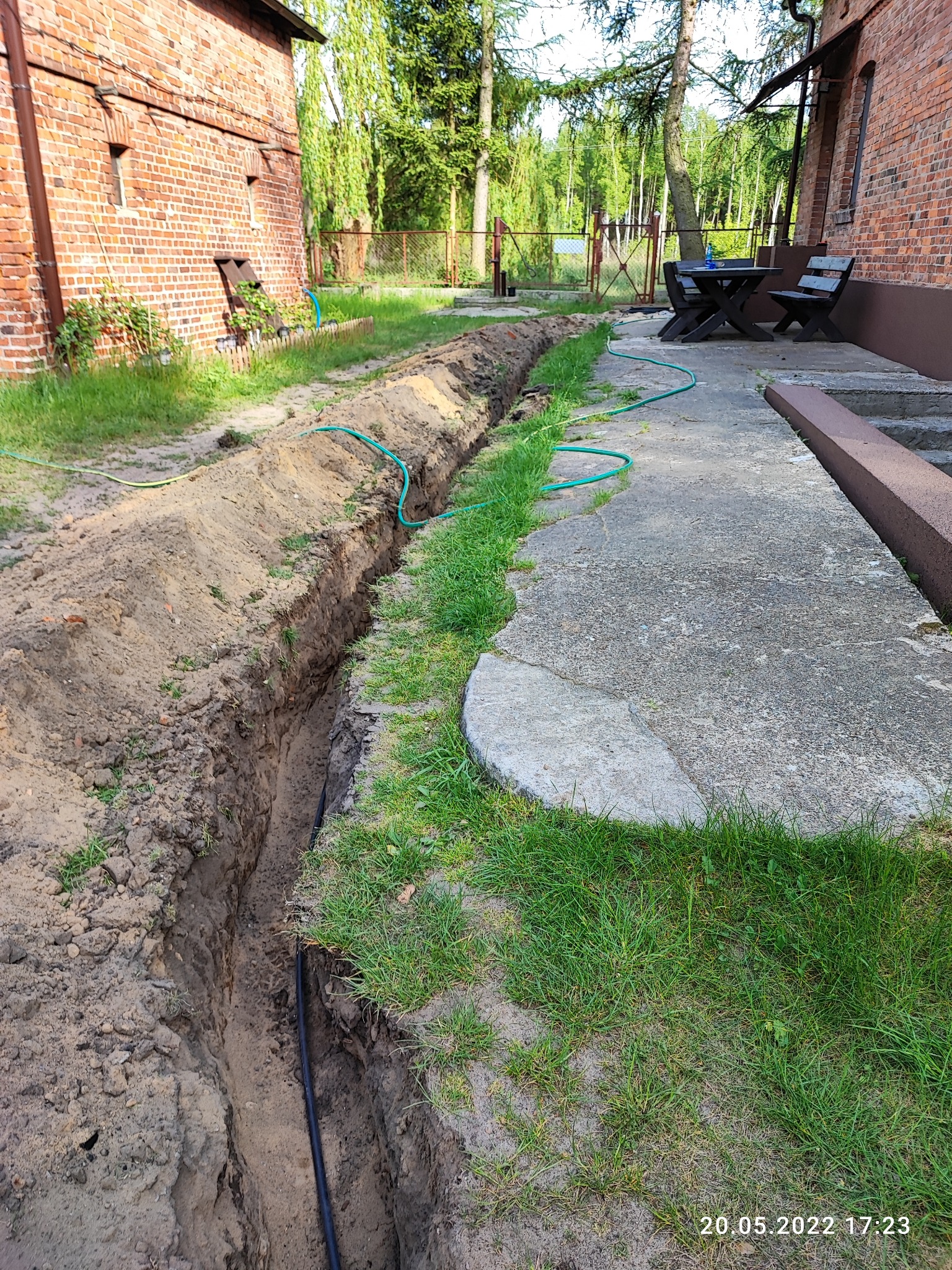 Wykopany wąski rów w ziemi, widoczna czarna rura na dnie, obok trawnik i betonowy chodnik, w tle budynek z czerwonej cegły i drzewa.