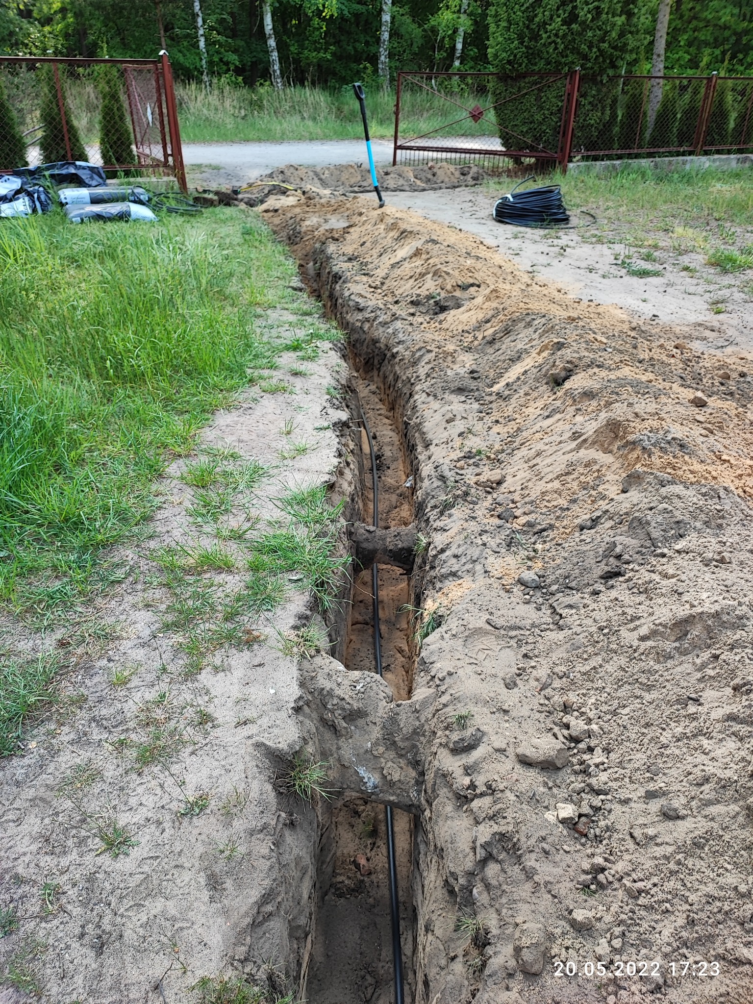 Wykopany rów w ziemi z widocznymi czarnymi rurami w środku, obok leżą worki i zwój kabli, w tle brama i zieleń.