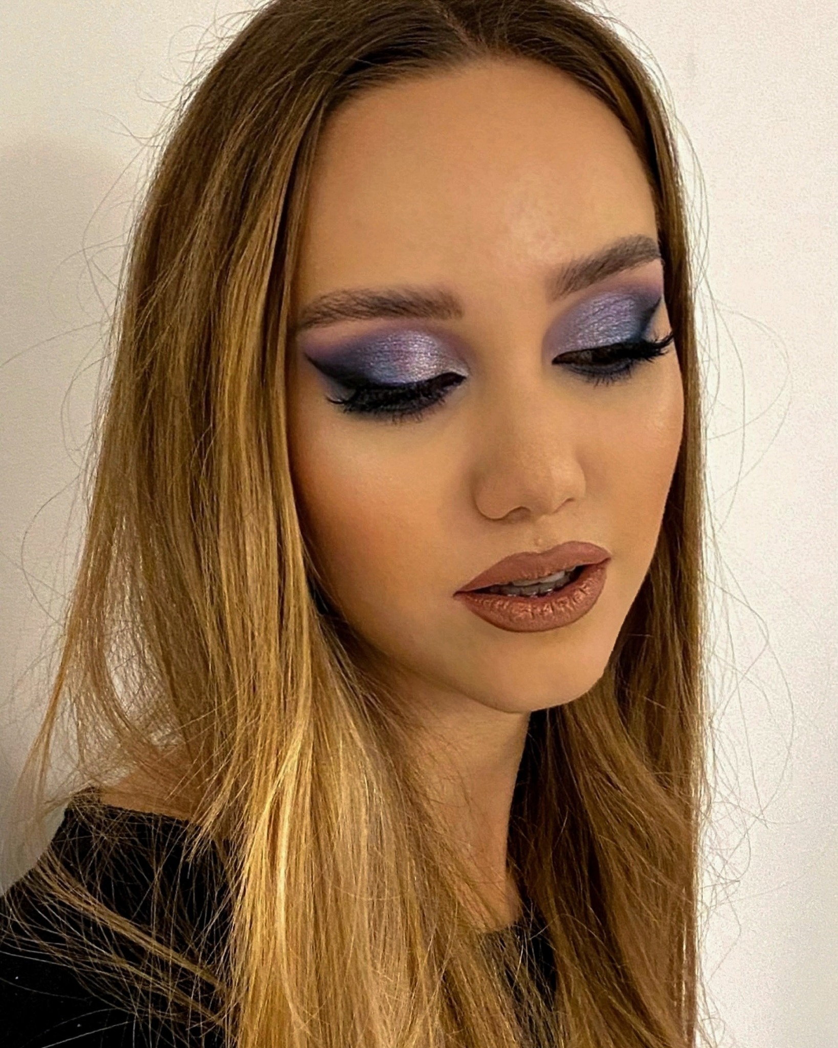 Portret kobiety z fioletowo-srebrnym cieniem na powiekach, czarną kreską i brązową pomadką, z rozpuszczonymi, jasnobrązowymi włosami sięgającymi ramion.
