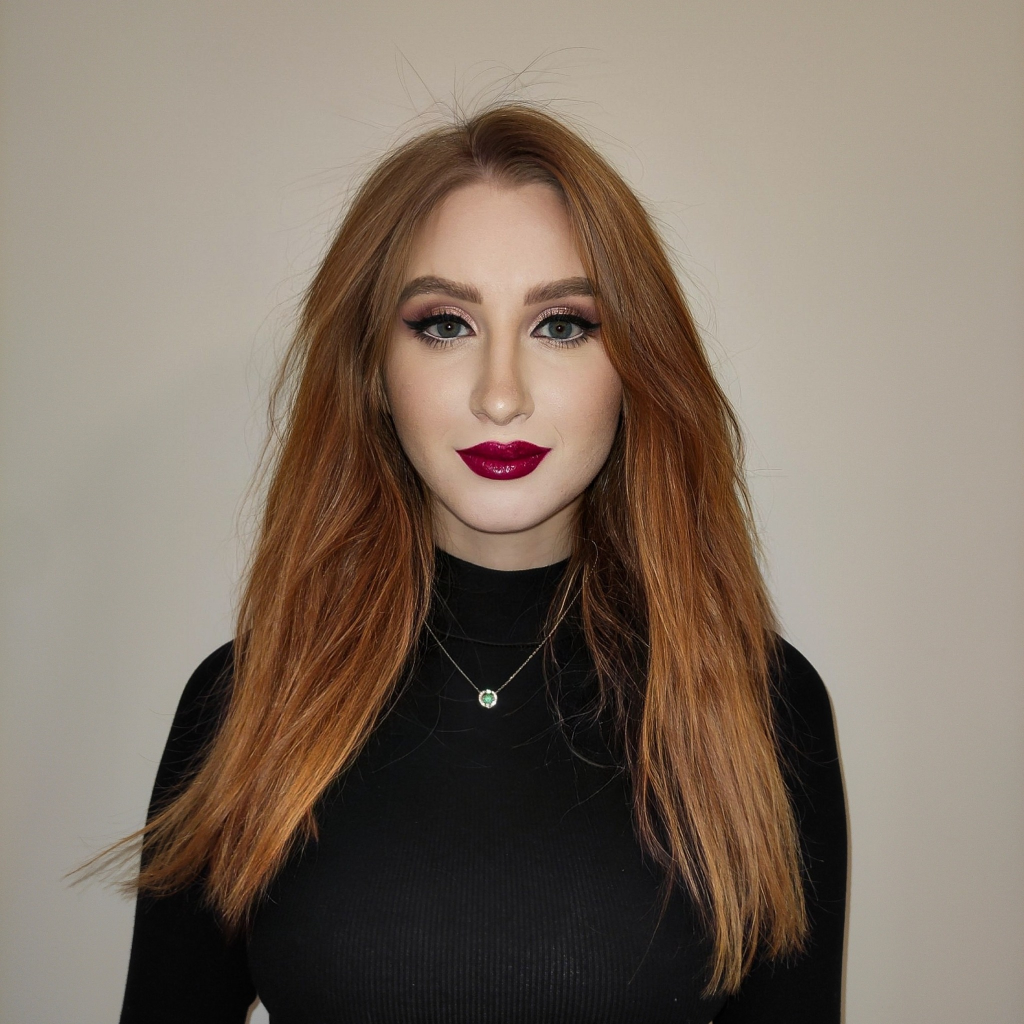 Portret kobiety o rudych włosach z intensywnym bordowym odcieniem szminki, podkreślonymi oczami z czarnym eyelinerem i delikatnym różowym cieniem, ubrana w czarny golf i naszyjnik z zielonym...