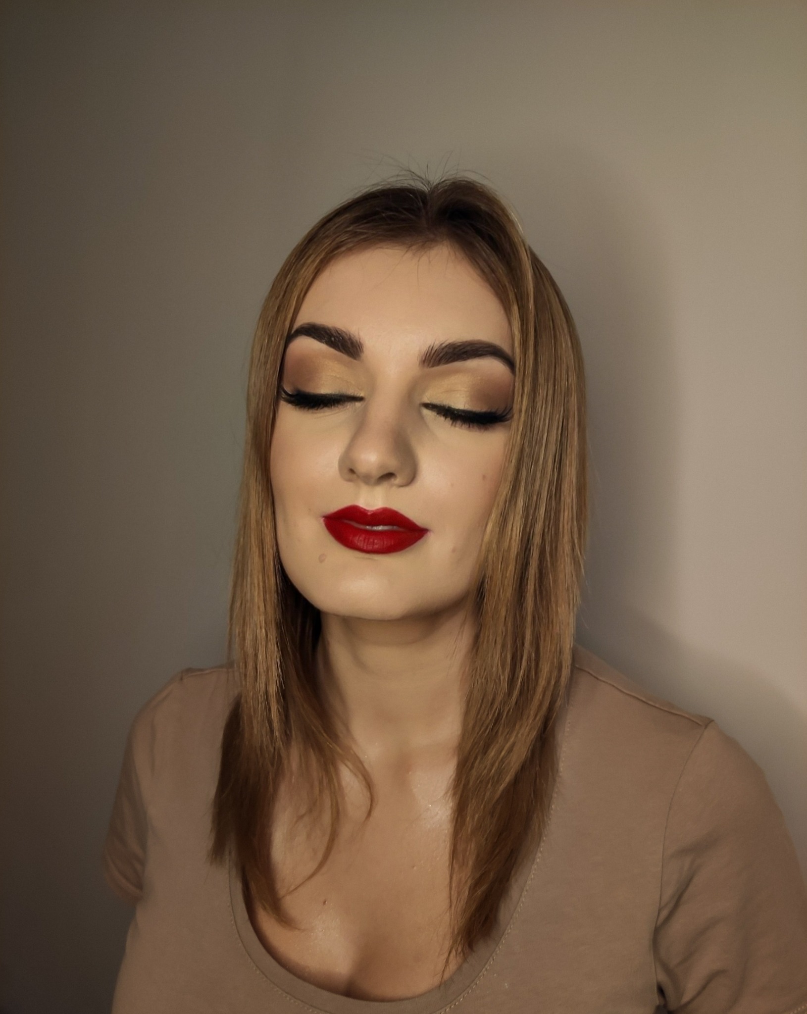 Portret kobiety z zamkniętymi oczami, prezentującej złoty cień na powiekach, długie sztuczne rzęsy i intensywnie czerwoną szminkę na ustach.