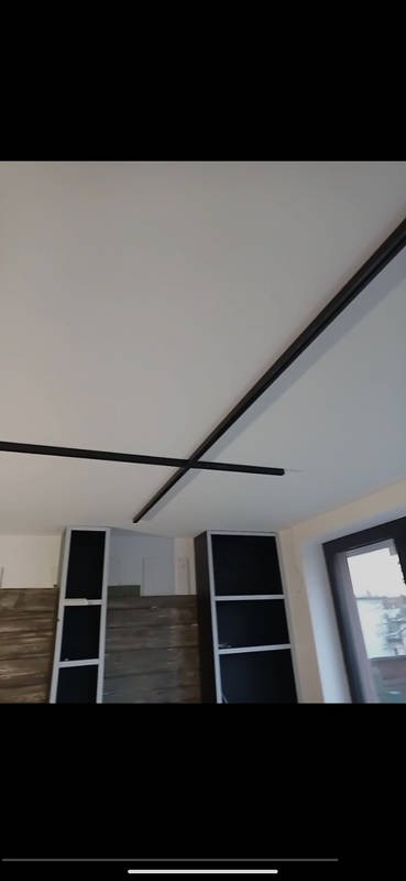 Nowoczesne oświetlenie sufitowe z czarnymi listwami LED ułożonymi na krzyż w jasnym wnętrzu z zabudową meblową i oknem.