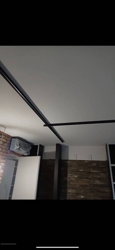 Czarna szyna oświetleniowa zamontowana na białym suficie w surowym wnętrzu, widoczna ceglana ściana i elementy zabudowy meblowej.