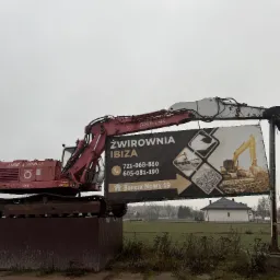 Czerwona koparka gąsienicowa na metalowej platformie z reklamą żwirowni 'IBIZA' na tle pochmurnego nieba i wiejskiego krajobrazu.