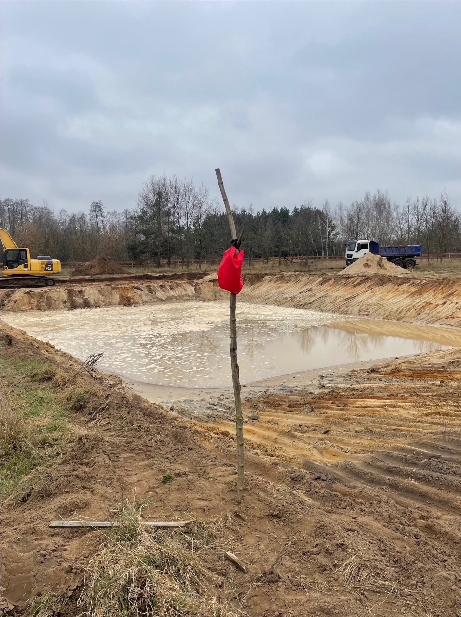 Wykopany staw wypełniony wodą, z żółtą koparką i ciężarówką w tle, oznaczony czerwoną torbą na tyczce.