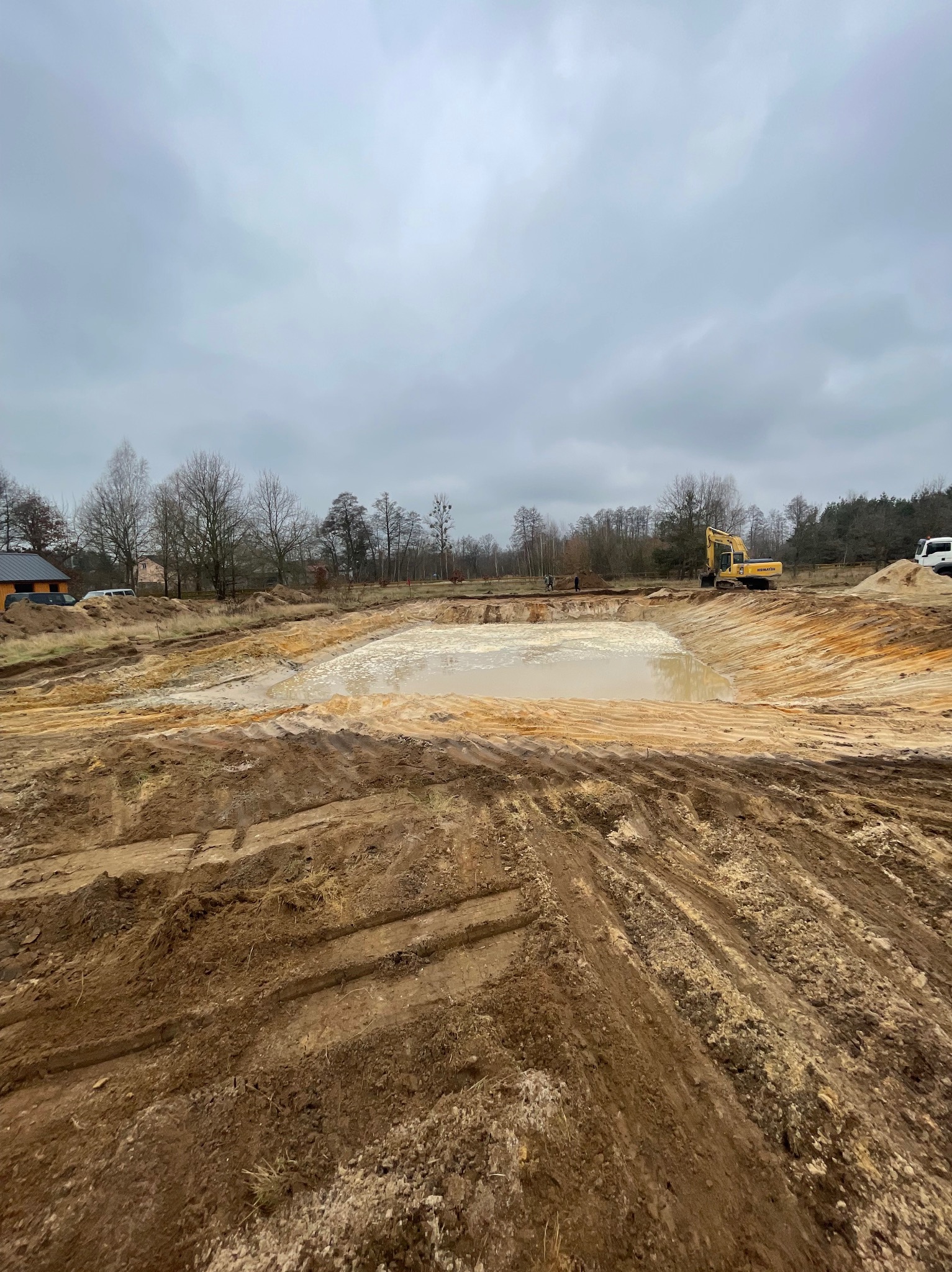 Żółta koparka Komatsu pracuje na skraju wykopu wypełnionego brudną wodą, widoczne ślady opon na ziemi, w tle las i pochmurne niebo.