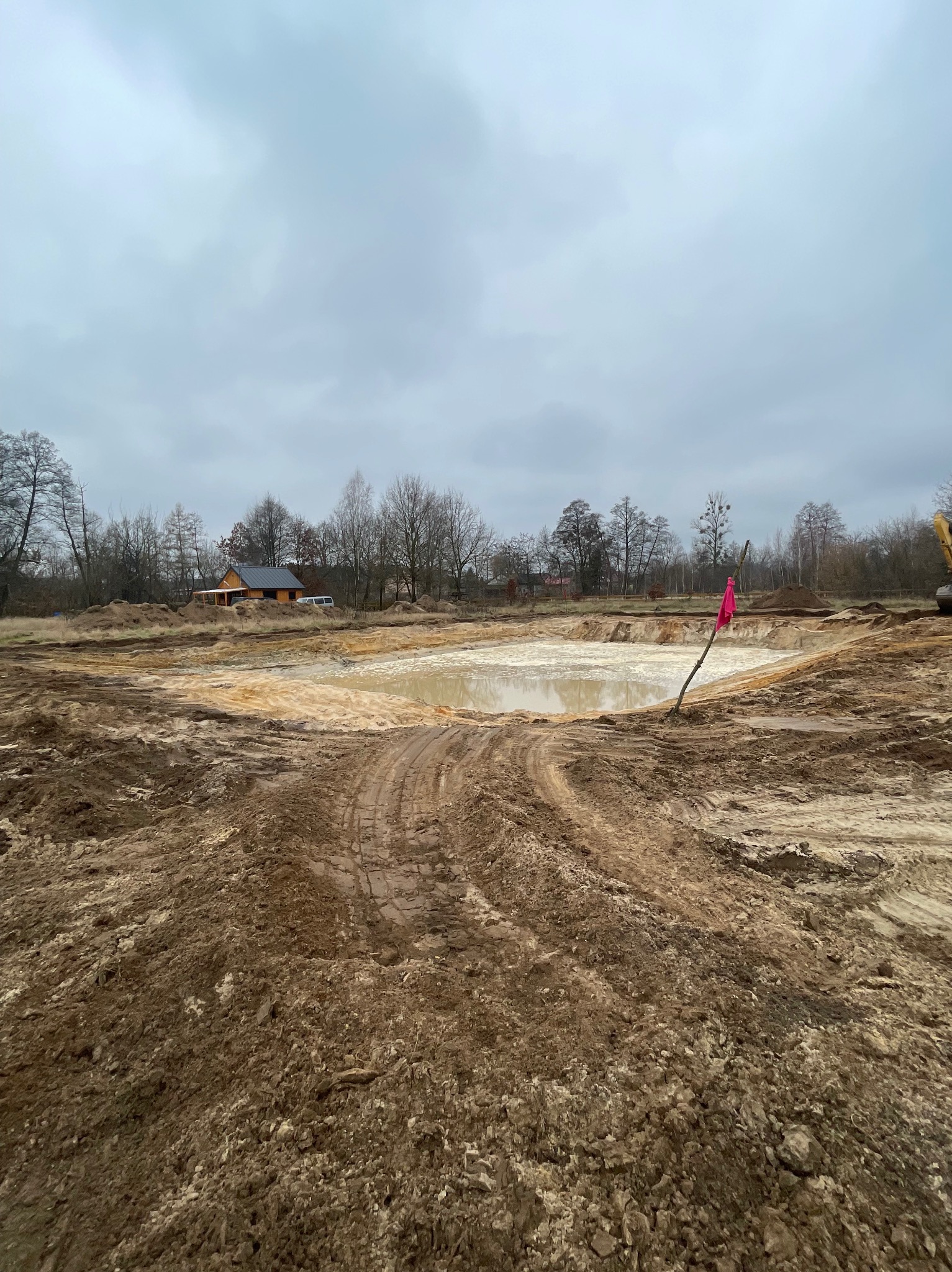 Wyrobisko wypełnione wodą, otoczone ziemią z wyraźnymi śladami opon, w tle widoczny budynek i drzewa pod pochmurnym niebem, z różową flagą na patyku.