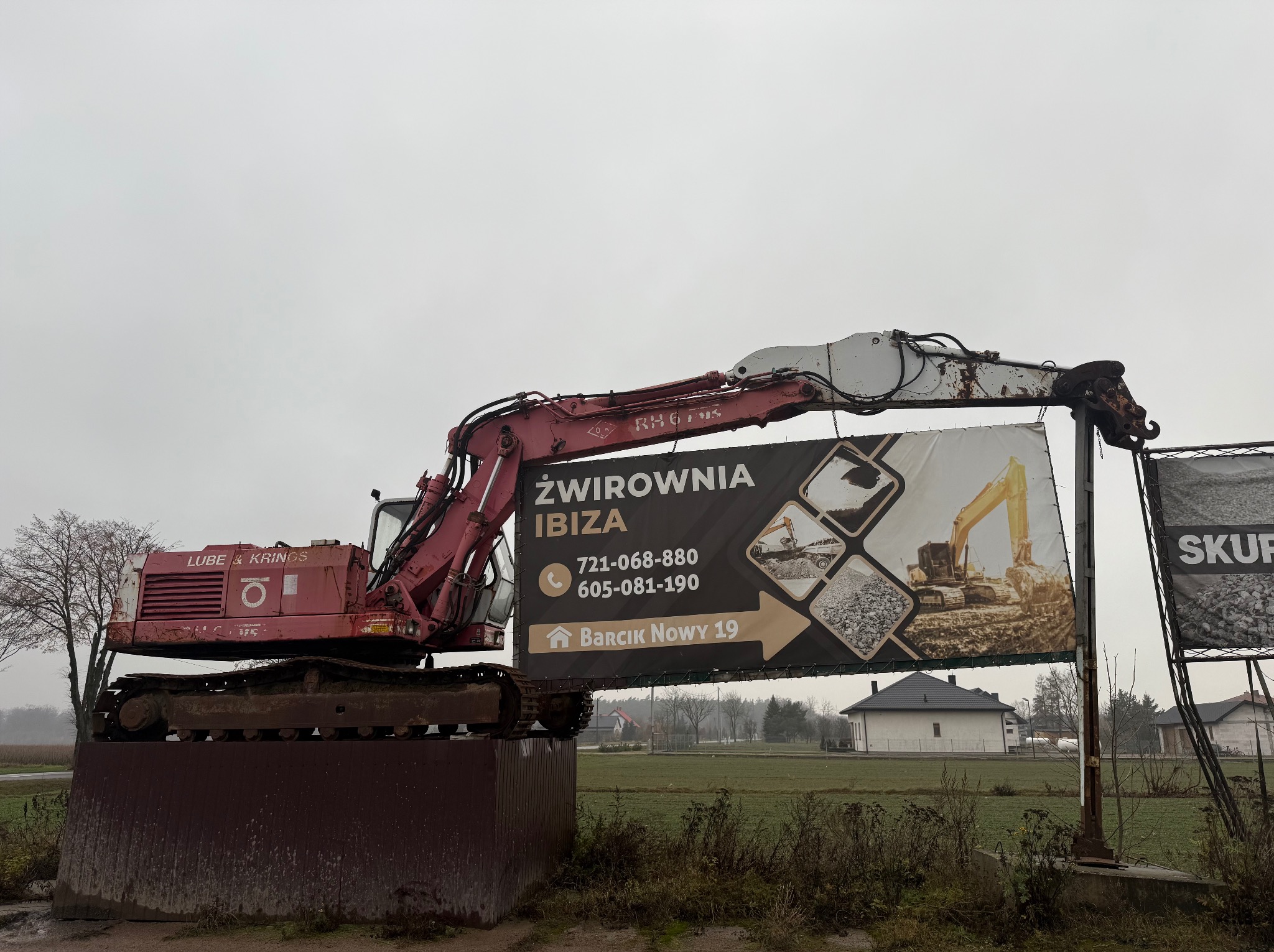 Czerwona koparka gąsienicowa na metalowej platformie z reklamą żwirowni 'IBIZA' na tle pochmurnego nieba i wiejskiego krajobrazu.