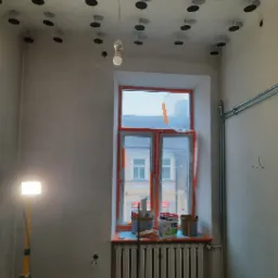 Przygotowanie do prac wykończeniowych w pokoju: surowa ściana, zabezpieczone okno folią, widoczne puszki z farbą na parapecie, lampa budowlana na statywie i zamontowane elementy do podwieszanego...