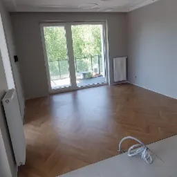 Wnętrze pokoju po remoncie: parkiet ułożony w jodełkę, świeżo pomalowane szare ściany, białe płytki podłogowe przy wejściu, grzejnik i okno balkonowe z widokiem na zieleń.