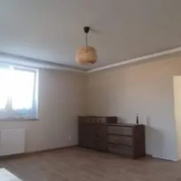 Pokój po odświeżeniu ścian w odcieniach beżu, z widoczną listwą sufitową i drewnianą podłogą; w tle otwarte drzwi do kolejnego pomieszczenia z widocznym bałaganem.