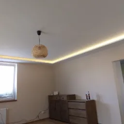 Jasne wnętrze pokoju z kremowymi ścianami, drewnianymi meblami i oświetleniem LED wzdłuż sufitu, uzupełnione wiszącą lampą z wikliny.