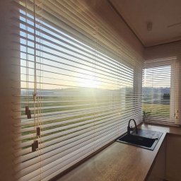 FHU ROYAL JACEK KOWALCZYK ROBERT KOWALCZYK SC - Jasne, drewniane żaluzje poziome w kuchni z widokiem na poranne słońce. Minimalistyczny design, ciepłe światło, nowoczesna armatura kuchenna.