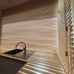 Beżowe żaluzje aluminiowe w kuchni, z widocznym zlewozmywakiem i padającym światłem słonecznym tworzącym cienie na blacie. Delikatne oświetlenie wnętrza.