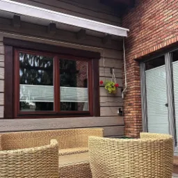 Białe plisy okienne zamontowane w brązowych oknach drewnianego domu z ceglanym fragmentem elewacji i rattanowymi meblami ogrodowymi.