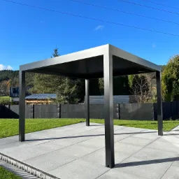 Nowoczesna, minimalistyczna pergola w kolorze antracytowym na tarasie z szarych płyt, otoczona zielonym trawnikiem i ogrodzeniem z siatki.