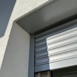 Fragment elewacji budynku z zamontowaną szarą, częściowo opuszczoną osłoną okienną z widocznym cieniem na ścianie.