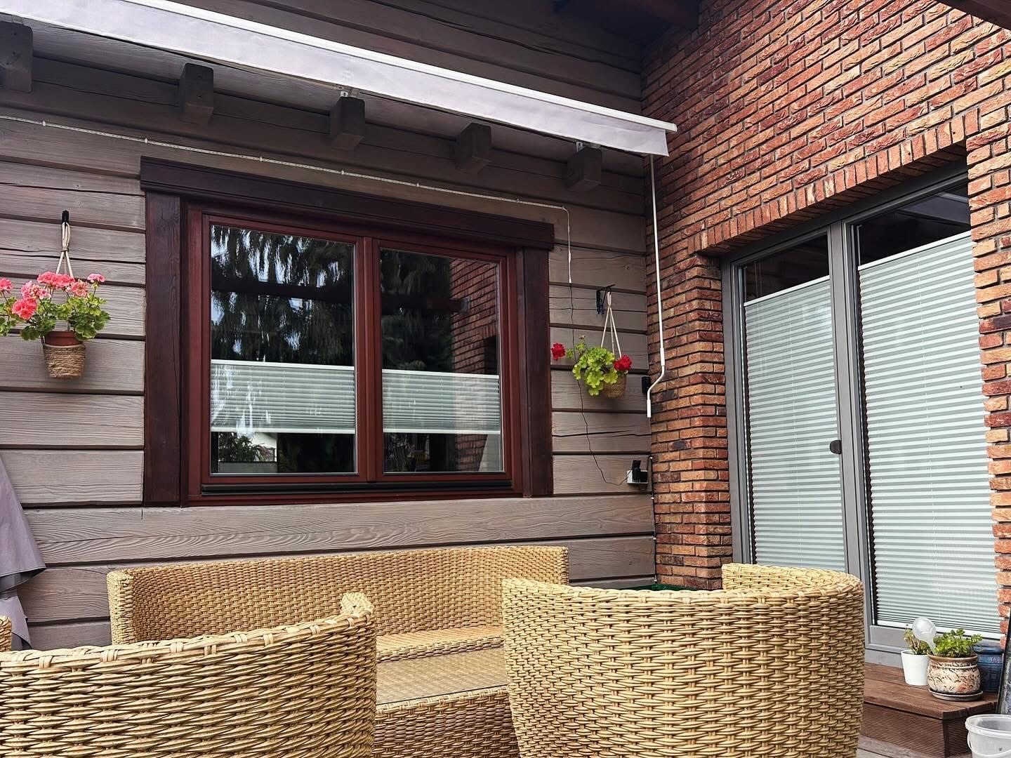 Białe plisy okienne zamontowane w brązowych oknach drewnianego domu z ceglanym fragmentem elewacji i rattanowymi meblami ogrodowymi.