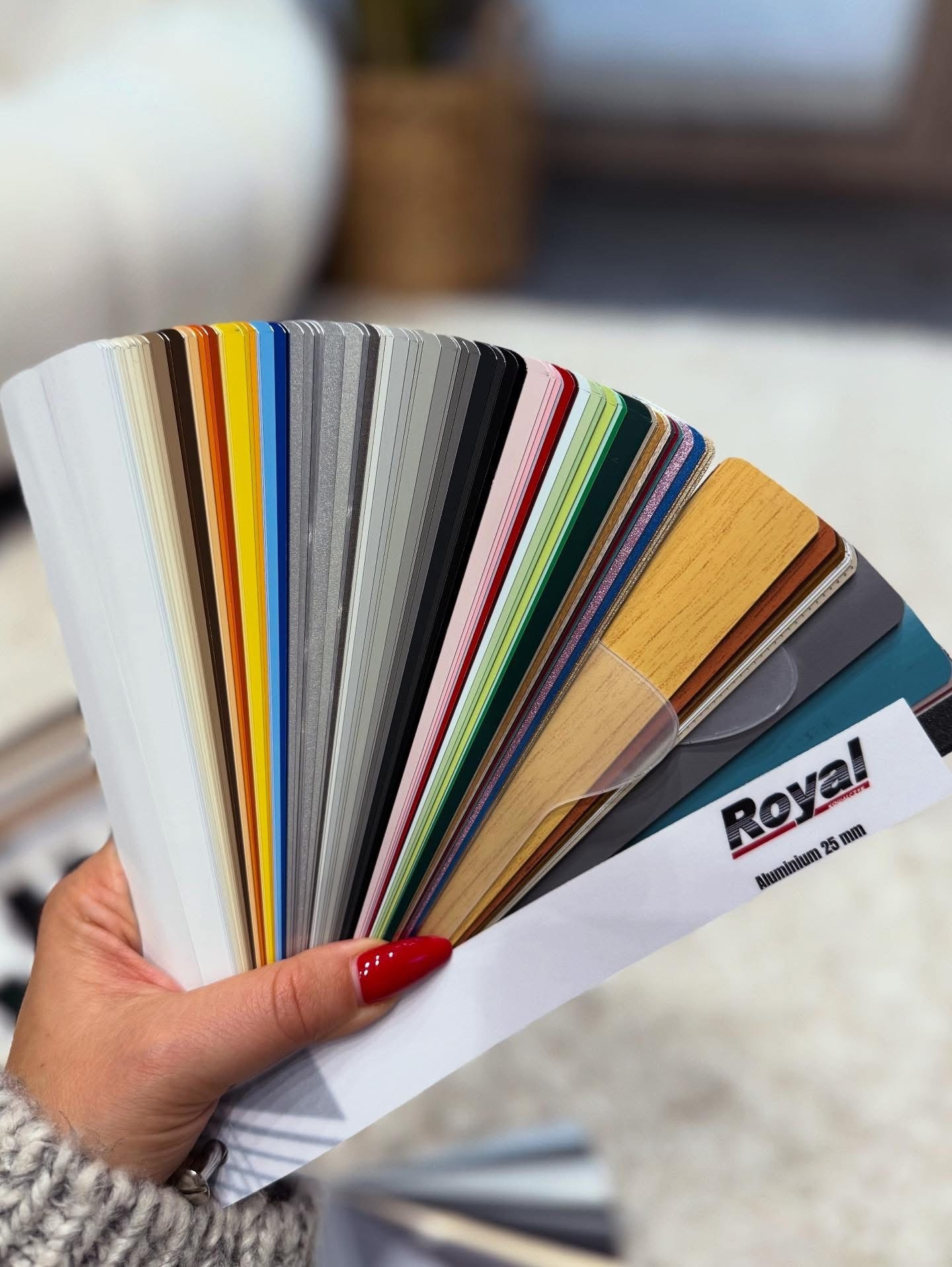 Paleta kolorów i faktur, w tym imitacji drewna, dla aluminiowych komponentów okiennych trzymana w dłoni, z widocznym logo 'Royal'.