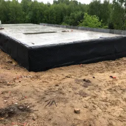 Wodoodporna izolacja fundamentów z folią kubełkową, widok na mokrą betonową płytę fundamentową i otaczający teren budowy