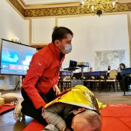 Instruktor w czerwonej kurtce klęczy obok osoby leżącej na czerwonej macie, przykrytej złotą folią termiczną, w tle sala szkoleniowa z projektorem i uczestnikami.