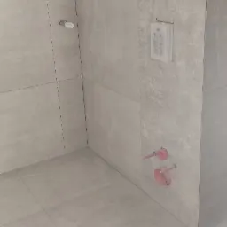 Świeżo ułożone, duże, szare płytki podłogowe i ścienne w łazience, widoczne krzyżyki dystansowe, instalacja podtynkowa i przyłącza wodne w kolorze różowym.