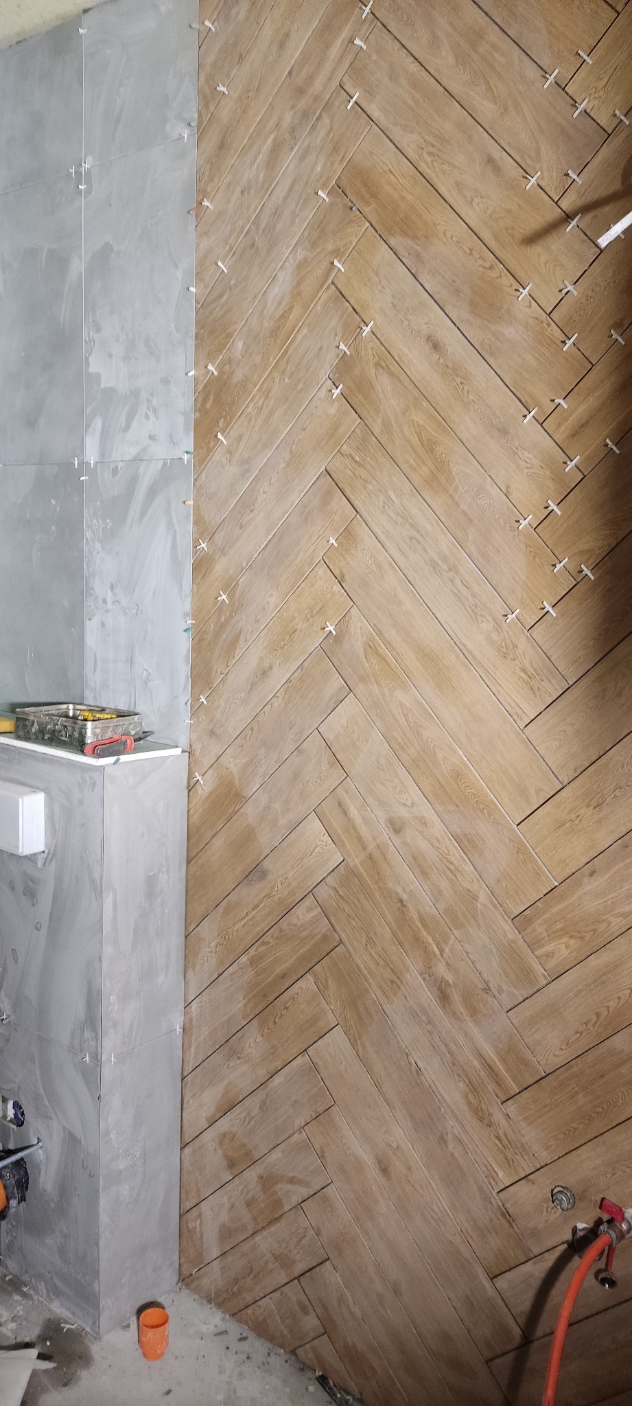 Układanie drewnopodobnych płytek w jodełkę na ścianie łazienki, widoczne krzyżyki dystansowe i fragment ściany z szarych płytek oraz narzędzia w tle.