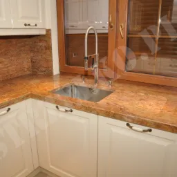 Kuchnia z blatem z kamienia w odcieniach brązu i beżu, kremowe szafki z ozdobnymi uchwytami, bateria kuchenna ze stali nierdzewnej, okno w drewnianej ramie.